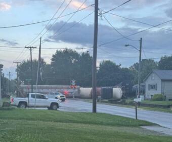 A freight train crosses a suburban street at a railroad crossing as vehicles wait; tanker railcars extend along the tracks under a cloudy sky.