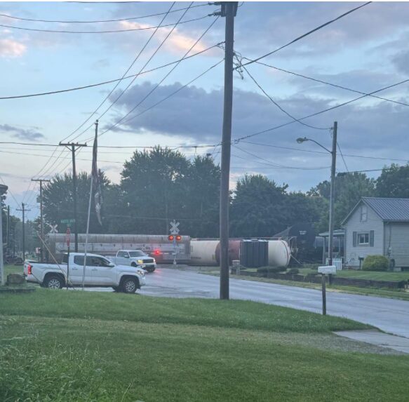 A freight train crosses a suburban street at a railroad crossing as vehicles wait; tanker railcars extend along the tracks under a cloudy sky.